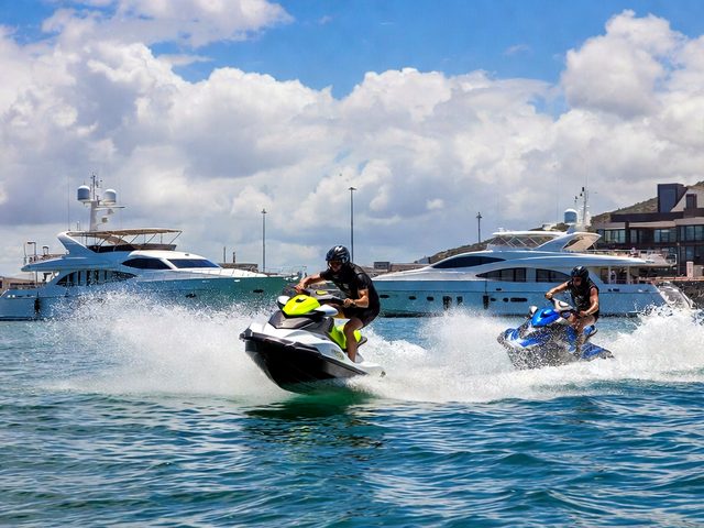 Two jet ski riders throwing spray in a marina with luxury yachts behind