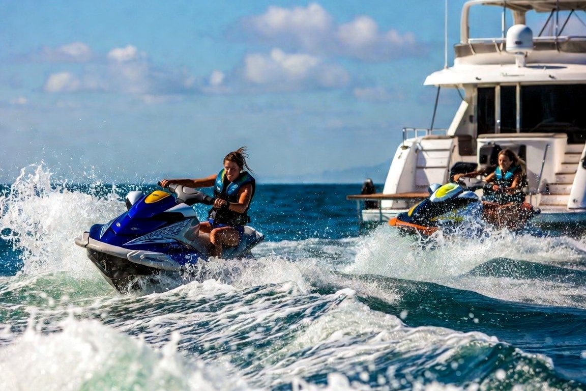 Two riders in life jackets launching jet skis off a yacht's swim platform