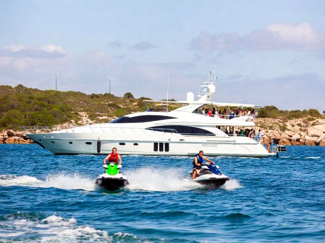 Two jet skis riding alongside a luxury motor yacht off a rocky coast